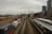 A bustling railway and shipping container yard set against an urban backdrop of tall buildings. Numerous colorful shipping containers are aligned on multiple train tracks, while a passenger train is stationed at a nearby platform. The sky is overcast, adding a dramatic tone to the scene.