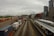 A bustling railway and shipping container yard set against an urban backdrop of tall buildings. Numerous colorful shipping containers are aligned on multiple train tracks, while a passenger train is stationed at a nearby platform. The sky is overcast, adding a dramatic tone to the scene.