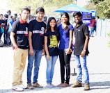 A group of five young adults stand in a sunny outdoor setting. They are smiling and appear relaxed. Three of them are wearing black T-shirts with text, while the others wear casual attire. There is a crowd and signs in the background, indicating a public event or gathering.