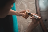 A person holds a vape device in one hand and a bottle of e-liquid labeled 'House Blend Premium E-liquid' in the other. The setting includes a soft beige surface and a glimpse of a rounded table edge, suggesting an indoor environment.