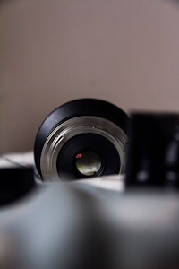 A close-up view of a camera lens with the focus on the front element. The surrounding environment is slightly blurred, highlighting the central lens. There are additional blurred objects in the foreground, likely other camera equipment.