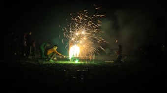 A person crouching on the ground lights a firework, causing bright sparks and smoke to illuminate the surroundings. Several figures stand in the background observing the display in a dark environment, possibly a beach or open field.