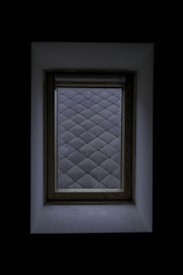 A window with a wooden frame set against a dark background. The view through the window displays a pattern of frosted glass squares, creating a diamond grid effect. The surrounding area is shadowed, highlighting the contrast between the window and its surroundings.