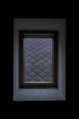A window with a wooden frame set against a dark background. The view through the window displays a pattern of frosted glass squares, creating a diamond grid effect. The surrounding area is shadowed, highlighting the contrast between the window and its surroundings.