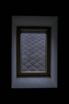A window with a wooden frame set against a dark background. The view through the window displays a pattern of frosted glass squares, creating a diamond grid effect. The surrounding area is shadowed, highlighting the contrast between the window and its surroundings.