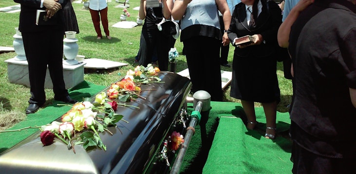 A group of people dressed in formal attire stand around a casket adorned with colorful roses. The setting appears to be a cemetery, with gravestones and manicured grass visible. Some individuals are holding books, likely hymnals or religious texts.