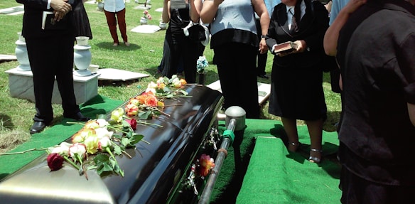 A group of people dressed in formal attire stand around a casket adorned with colorful roses. The setting appears to be a cemetery, with gravestones and manicured grass visible. Some individuals are holding books, likely hymnals or religious texts.