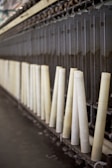 A row of large, vertical spinning spindles, likely part of a textile or yarn production machine. The spindles are light-colored and arranged neatly along a metal framework.