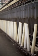 A row of large, vertical spinning spindles, likely part of a textile or yarn production machine. The spindles are light-colored and arranged neatly along a metal framework.