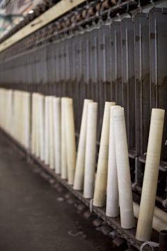A row of large, vertical spinning spindles, likely part of a textile or yarn production machine. The spindles are light-colored and arranged neatly along a metal framework.