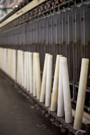 A row of large, vertical spinning spindles, likely part of a textile or yarn production machine. The spindles are light-colored and arranged neatly along a metal framework.