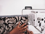 A person's hand is guiding patterned fabric through a sewing machine. The fabric features a detailed floral design with beige and brown leaves against a black background. The sewing machine is white with visible dials and mechanisms.