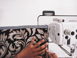 A person's hand is guiding patterned fabric through a sewing machine. The fabric features a detailed floral design with beige and brown leaves against a black background. The sewing machine is white with visible dials and mechanisms.