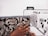 A person's hand is guiding patterned fabric through a sewing machine. The fabric features a detailed floral design with beige and brown leaves against a black background. The sewing machine is white with visible dials and mechanisms.
