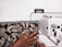 A person's hand is guiding patterned fabric through a sewing machine. The fabric features a detailed floral design with beige and brown leaves against a black background. The sewing machine is white with visible dials and mechanisms.