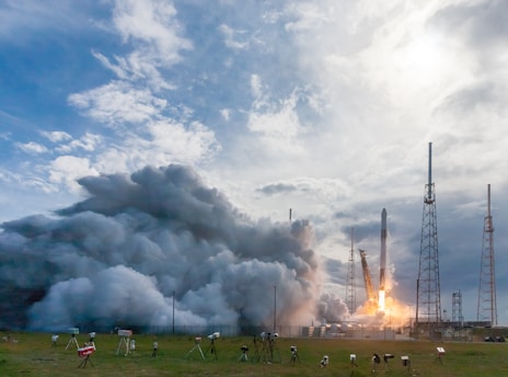A rocket is being launched amidst large billows of smoke with clear blue sky and scattered clouds above. Several towers and structures are visible around the launch site, and multiple cameras are set up on the ground to capture the event.