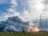 A rocket is being launched amidst large billows of smoke with clear blue sky and scattered clouds above. Several towers and structures are visible around the launch site, and multiple cameras are set up on the ground to capture the event.