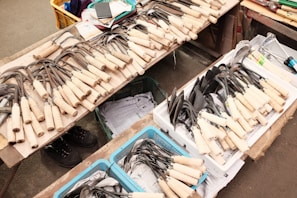 A variety of gardening tools with wooden handles are neatly arranged on a table. The tools include different kinds of hoes and spades, some in metal baskets or trays. Nearby, there are cardboard boxes and other miscellaneous items like a notebook and a smartphone. The setting appears to be a market or shop floor with visible concrete or tile flooring.