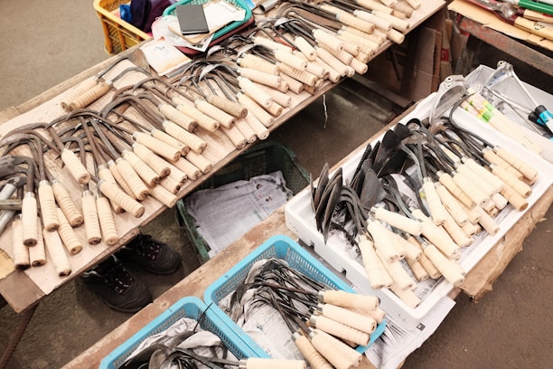 A variety of gardening tools with wooden handles are neatly arranged on a table. The tools include different kinds of hoes and spades, some in metal baskets or trays. Nearby, there are cardboard boxes and other miscellaneous items like a notebook and a smartphone. The setting appears to be a market or shop floor with visible concrete or tile flooring.