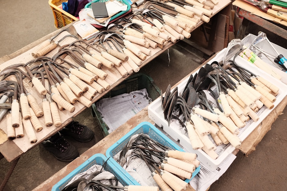 A variety of gardening tools with wooden handles are neatly arranged on a table. The tools include different kinds of hoes and spades, some in metal baskets or trays. Nearby, there are cardboard boxes and other miscellaneous items like a notebook and a smartphone. The setting appears to be a market or shop floor with visible concrete or tile flooring.