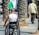 A person in a wheelchair is seen from behind on a paved sidewalk lined with palm trees. The person has a shaved head and is wearing a sleeveless white shirt, revealing a tattooed arm. Another person is walking ahead, wearing a light green shirt and dark pants.