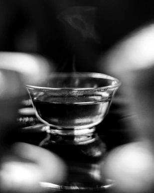 A steaming glass cup filled with a hot beverage is placed in focus, with a blurred background. Light reflections on the glass create a sense of depth and texture, while the steam rising adds a dynamic element.