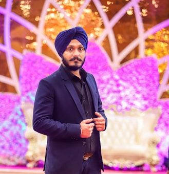 A man with a black beard wearing a dark suit and blue turban stands confidently in front of a vibrant and colorful backdrop, featuring purple and gold decorative elements. The scene is likely set at an event, providing a festive atmosphere.