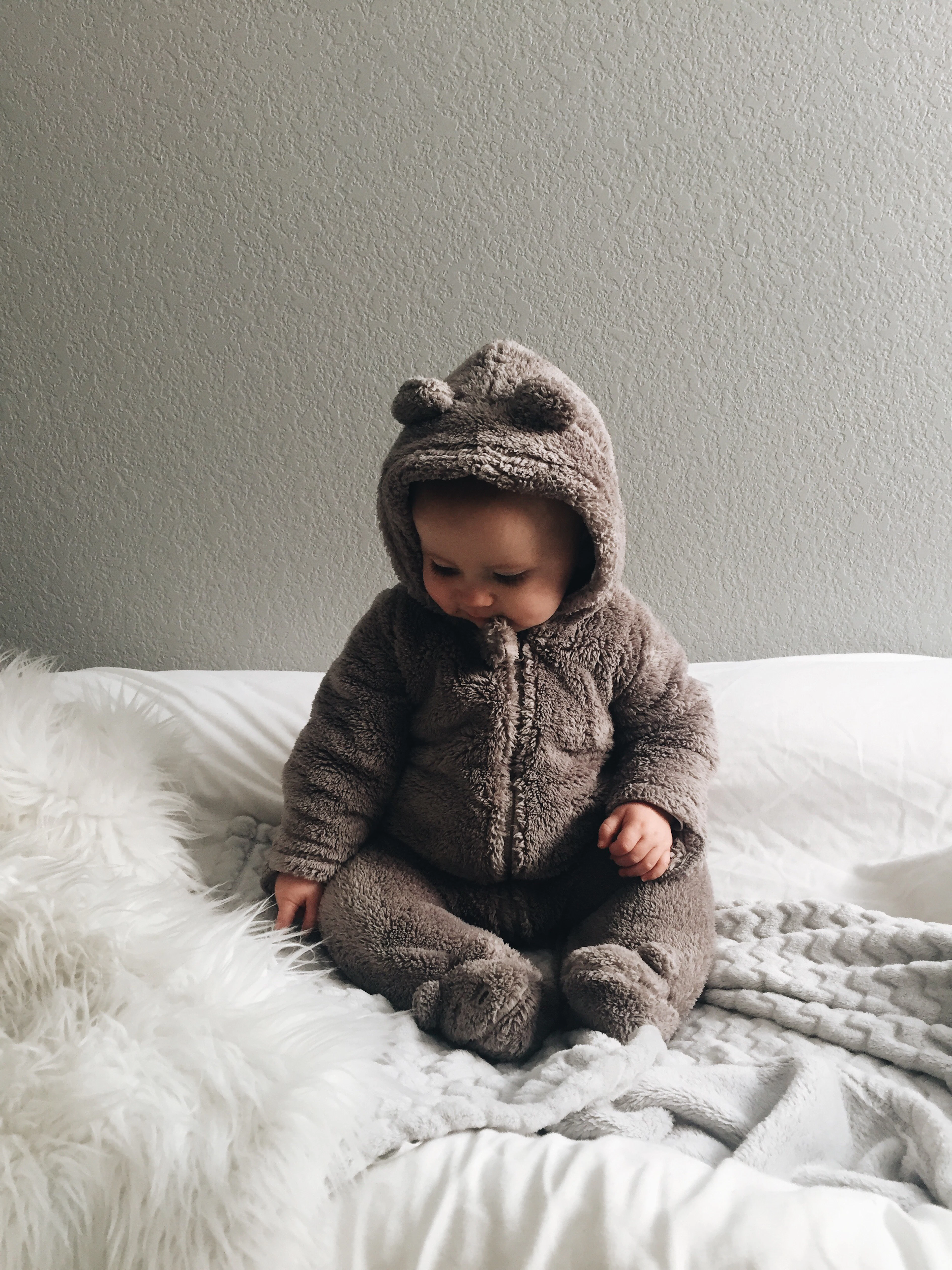 A baby in a soft bear suit sitting on white bedding