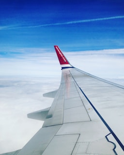 An airplane wing extends outward against a background of a blue sky and white clouds, with wispy contrails visible in the distance. The tip of the wing features a red accent with text, contrasting against the softer colors around it.