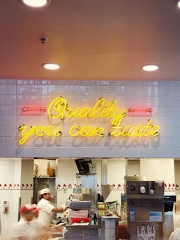 Neon sign in yellow text reading 'Quality you can taste' is mounted on a white tiled wall inside a fast food restaurant. Beneath the sign, several workers dressed in white uniforms and red hats are busy at a counter preparing food.