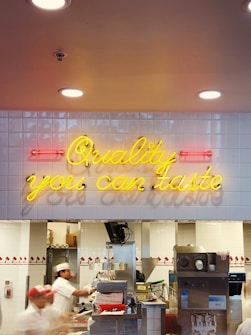 Neon sign in yellow text reading 'Quality you can taste' is mounted on a white tiled wall inside a fast food restaurant. Beneath the sign, several workers dressed in white uniforms and red hats are busy at a counter preparing food.