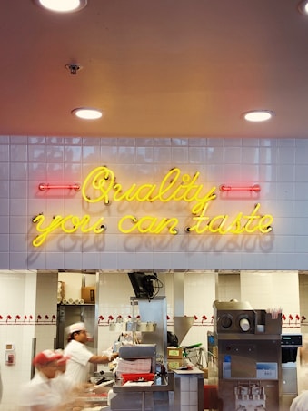 Neon sign in yellow text reading 'Quality you can taste' is mounted on a white tiled wall inside a fast food restaurant. Beneath the sign, several workers dressed in white uniforms and red hats are busy at a counter preparing food.