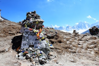 A stone memorial adorned with colorful prayer flags sits atop a rocky terrain with majestic snow-capped mountains in the background. The large stone bears inscriptions and is surrounded by smaller rocks. The sky is clear and blue, enhancing the serene natural setting.