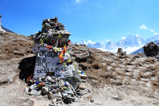 A stone memorial adorned with colorful prayer flags sits atop a rocky terrain with majestic snow-capped mountains in the background. The large stone bears inscriptions and is surrounded by smaller rocks. The sky is clear and blue, enhancing the serene natural setting.
