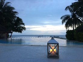 A tranquil beachfront setting featuring an infinity pool with calm water reflecting the sky. Tall palm trees frame the scene on both sides, and a single candle in a decorative lantern is placed in the foreground, adding warmth to the cool tones. The horizon meets a serene sea under a pale blue twilight sky.