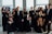 A group of people dressed formally in suits and dresses, posing confidently inside a well-lit room with large windows. Some individuals make playful gestures while others stand with arms crossed. The mood is lively, suggesting a celebratory event or gathering.