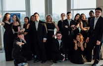 A group of people dressed formally in suits and dresses, posing confidently inside a well-lit room with large windows. Some individuals make playful gestures while others stand with arms crossed. The mood is lively, suggesting a celebratory event or gathering.