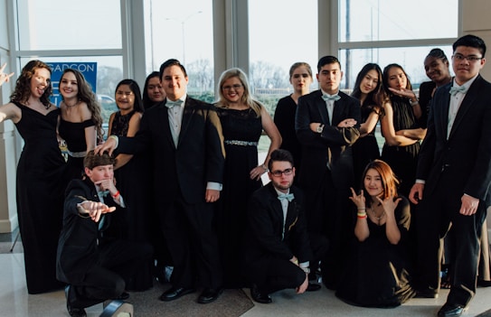 A group of people dressed formally in suits and dresses, posing confidently inside a well-lit room with large windows. Some individuals make playful gestures while others stand with arms crossed. The mood is lively, suggesting a celebratory event or gathering.
