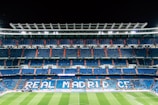 An expansive football stadium with empty blue seating areas primarily in the middle levels. The words 'REAL MADRID C.F.' are prominently displayed in large letters across the seats. The field is visible at the bottom, with a few scattered people on the pitch and more dispersed in the stands. Bright stadium lights illuminate the scene, contrasting with the dark night sky.