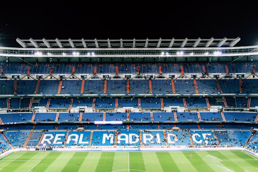An expansive football stadium with empty blue seating areas primarily in the middle levels. The words 'REAL MADRID C.F.' are prominently displayed in large letters across the seats. The field is visible at the bottom, with a few scattered people on the pitch and more dispersed in the stands. Bright stadium lights illuminate the scene, contrasting with the dark night sky.