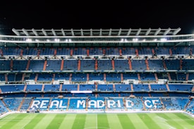 An expansive football stadium with empty blue seating areas primarily in the middle levels. The words 'REAL MADRID C.F.' are prominently displayed in large letters across the seats. The field is visible at the bottom, with a few scattered people on the pitch and more dispersed in the stands. Bright stadium lights illuminate the scene, contrasting with the dark night sky.