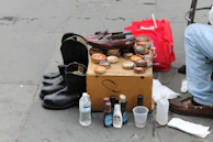A small street setup featuring shoe shining items. There are polished leather shoes and boots positioned in neat lines. Shoe polish tins, brushes, and other shoe care products are displayed on a cardboard box. A water bottle and other miscellaneous cleaning products are nearby. A person wearing jeans is partially visible, sitting beside the setup.