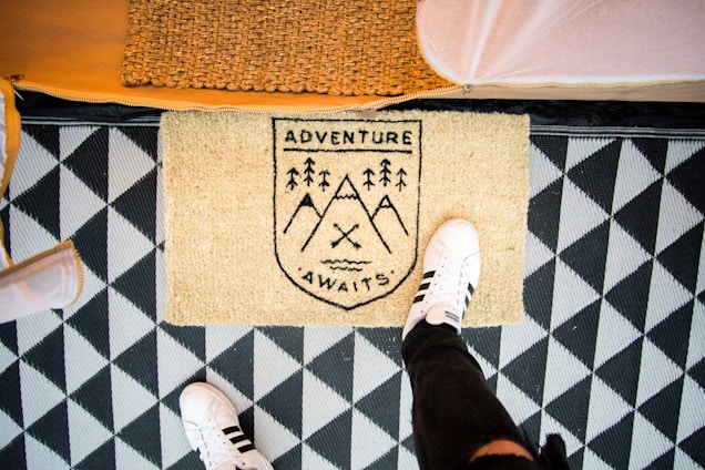 A doormat with the text 'Adventure Awaits' is placed on a geometric patterned floor. One foot wearing a white sneaker with black stripes stands partially on the mat. The background includes a woven basket and hints of surrounding structures.
