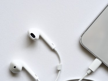 A pair of white wired earphones lies on a white surface, next to a smartphone partially visible with a connected charging cable. The minimalistic setup creates a clean and modern look.