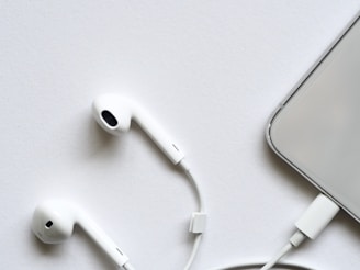 A pair of white wired earphones lies on a white surface, next to a smartphone partially visible with a connected charging cable. The minimalistic setup creates a clean and modern look.