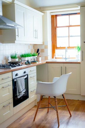 A modern kitchen with white cabinets and wooden countertops features a gas stovetop and a built-in oven. Green plants in white pots are placed on the countertop, adding a touch of nature. A white chair with wooden legs is positioned in front of the oven, and natural light pours in through a large window with a wooden frame, illuminating the space.