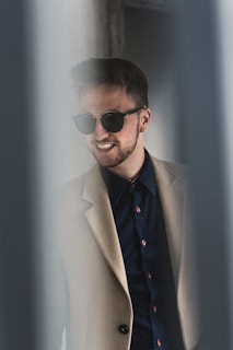 A man with short hair wears sunglasses and smiles while dressed in a beige coat over a dark shirt with red buttons. The image captures a stylish and casual look with a soft, out-of-focus foreground.