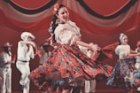 A group of dancers wearing traditional folk costumes perform energetically on stage. The central figure is a woman spinning in a red dress with floral patterns, holding a red shawl. Other dancers in similar attire appear blurred in the background, adding a sense of motion.