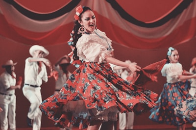 A group of dancers wearing traditional folk costumes perform energetically on stage. The central figure is a woman spinning in a red dress with floral patterns, holding a red shawl. Other dancers in similar attire appear blurred in the background, adding a sense of motion.