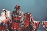 A woman is performing a traditional dance, wearing a vibrant, flowing dress adorned with floral patterns. Her hair is styled neatly with a decorative flower, and she is fully engaged in the graceful movement. The setting suggests a cultural or stage performance with other dancers partially visible in similar attire.